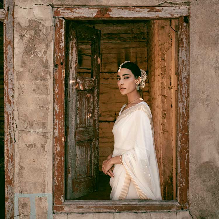 Woman in white traditional saree with floral jewelry standing in rustic doorway