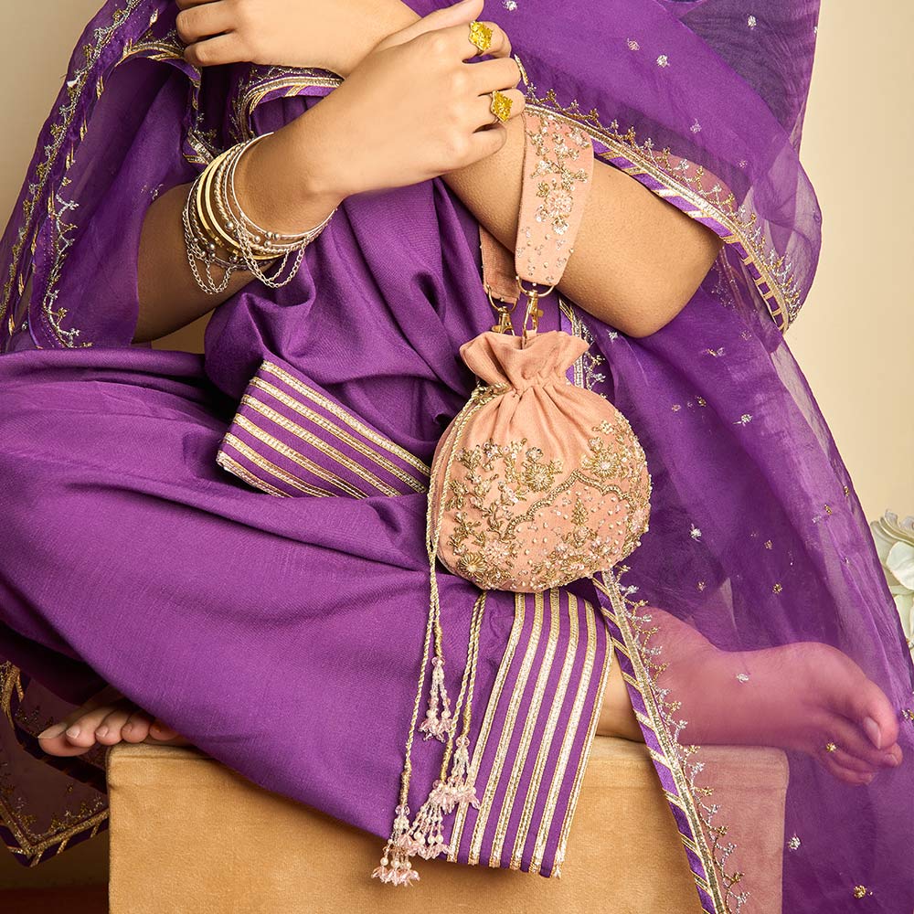 Woman in a purple embroidered saree holding a pink embellished potli bag, wearing gold bangles and rings.